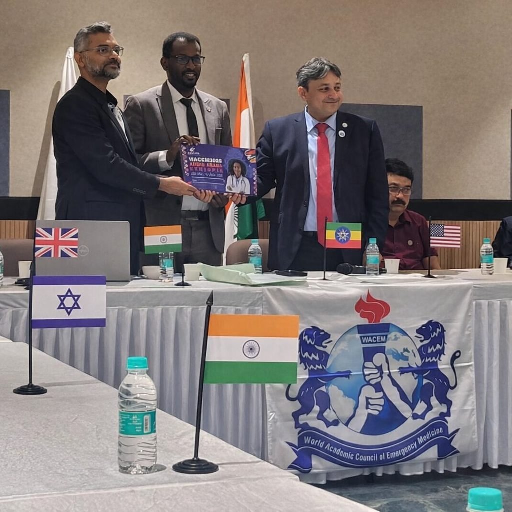 Dr. Woldesenbet Waganew, center, receives a purple WACEM 2026 Addis Ababa plaque from Dr. Vimal Krishnan, left, and Dr. Sagar Galwankar, right, at a table with the WACEM banner and small national flags.